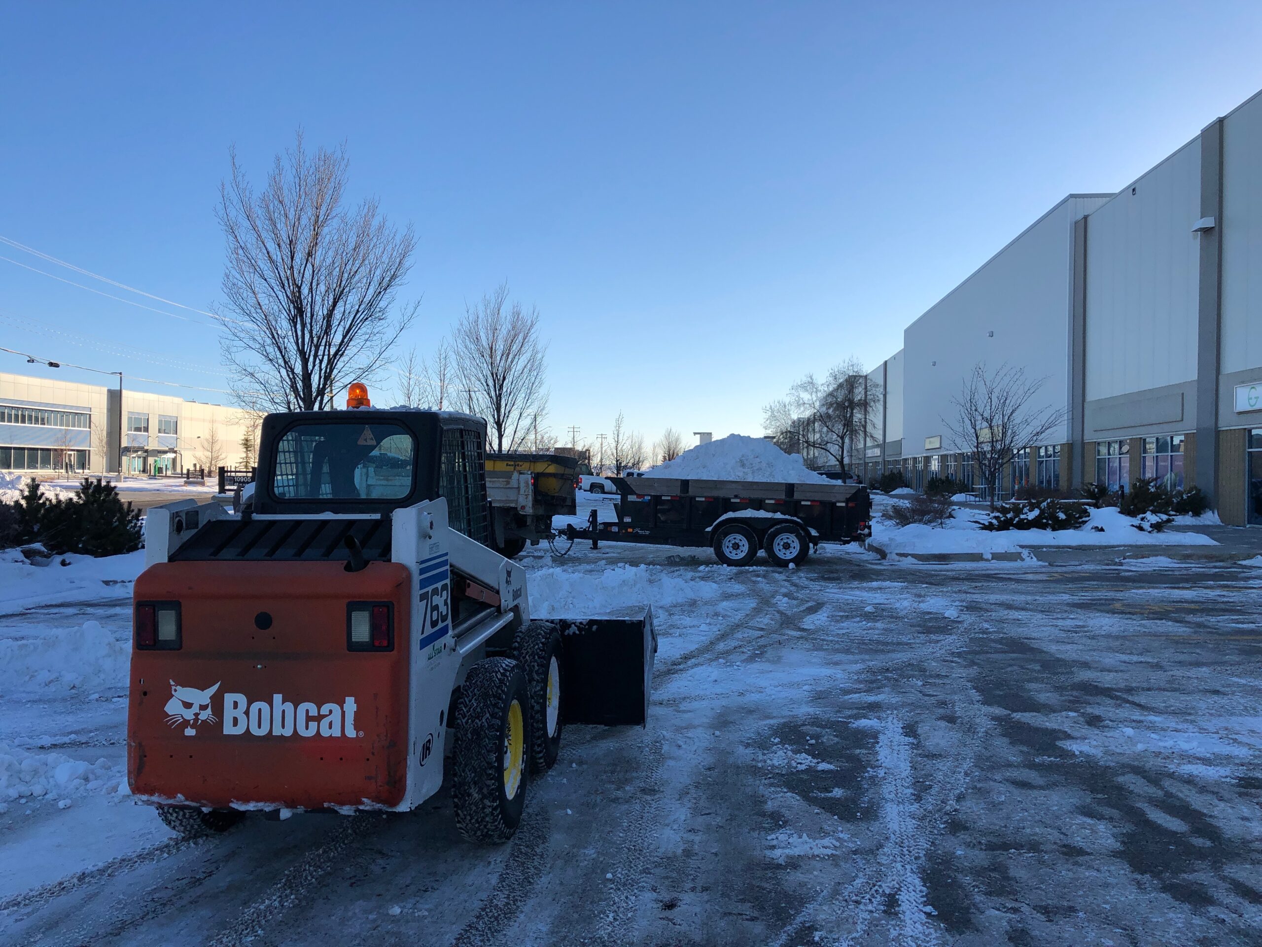 Bobcat skid steer and trailer clearing snow at an industrial building